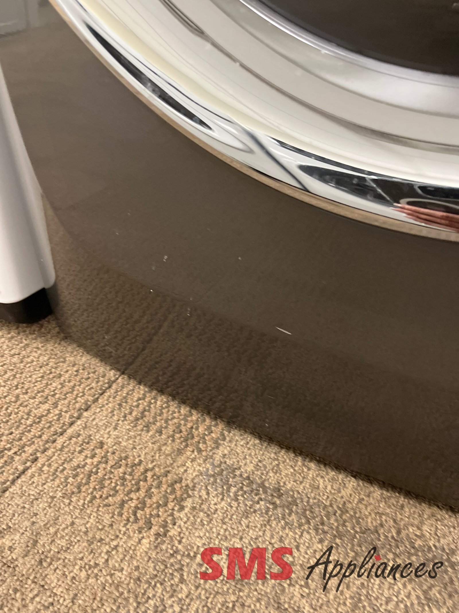 Refurbished-Dryers-EFMC527UTT-5-1-scaled-1.jpg Close-up of a front-loading washing machine with a shiny chrome drum rim on a beige carpet; SMS Appliances logo partially visible at bottom right.
